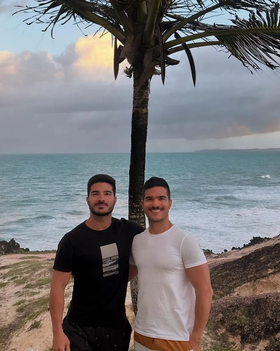 Os fundadores da Kaya Mundi lado a lado sob um coqueiro, num mirante à beira-mar, com o oceano e nuvens de fim de tarde ao fundo
